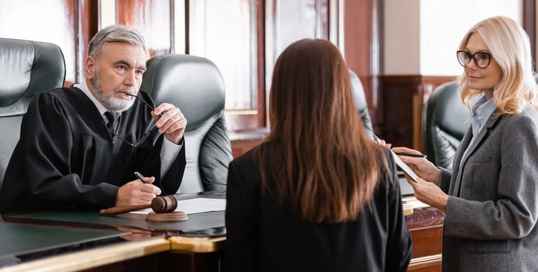 A judge sits at a courtroom bench, listening intently to two women standing before him. One woman holds papers and a pen, while the other stands with her back to the camera.