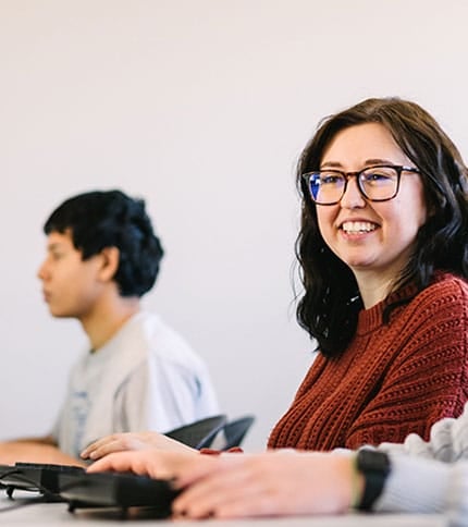 Two people seated at a desk in a classroom setting. The person in the foreground is smiling and wearing glasses and a red sweater. The second person, appearing focused, is seated further back, wearing a gray shirt.