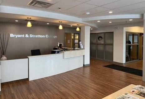 The reception area at the Henrietta NY campus of Bryant & Stratton College features a modern front desk, warm wood flooring, and soft overhead lighting.