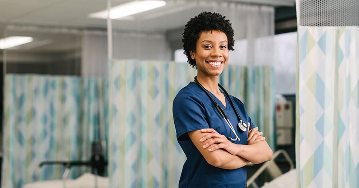 Smiling healthcare professional in blue scrubs, crossing arms, wearing a stethoscope, standing in a medical facility.