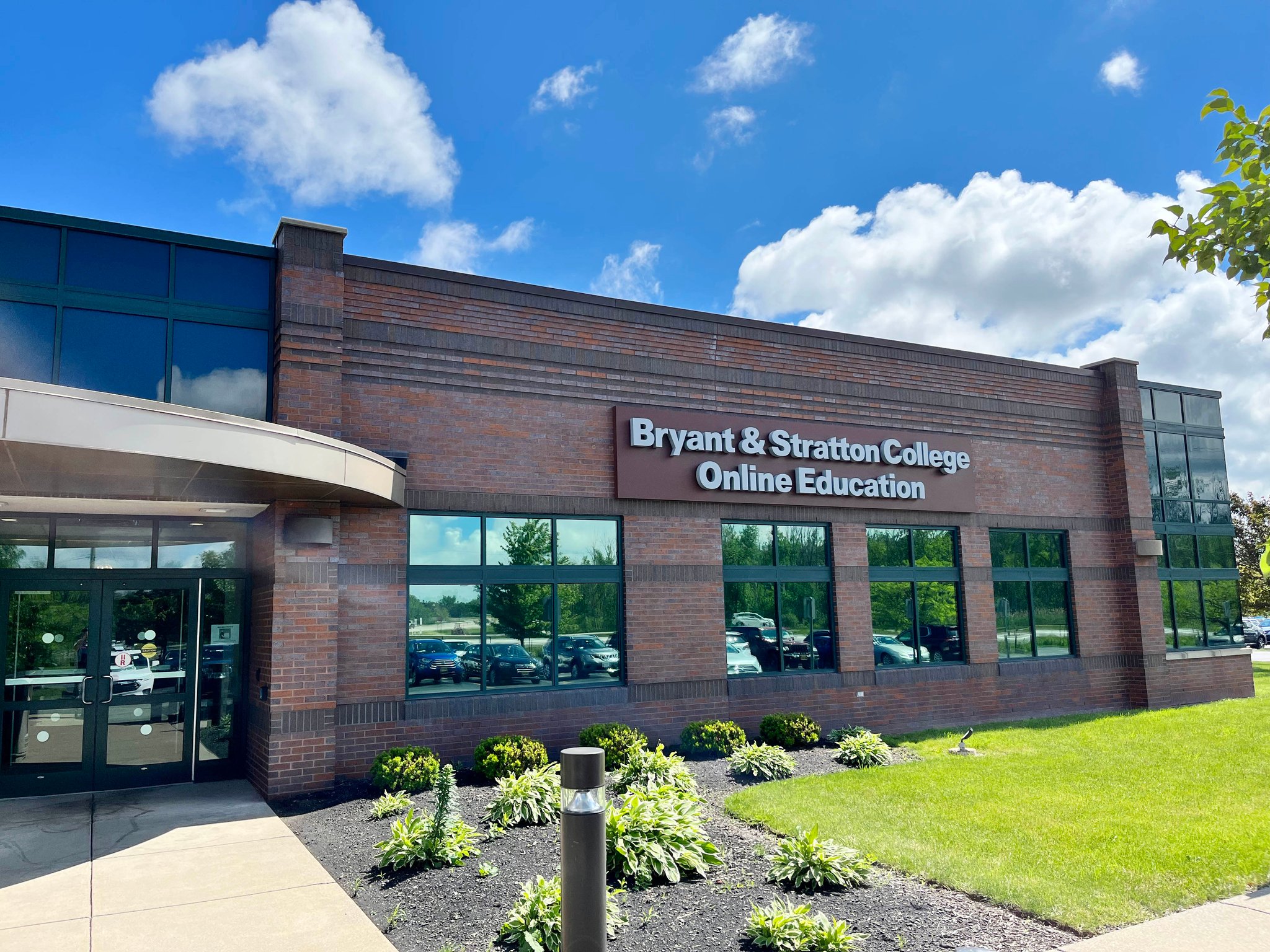 A brick building with a sign reading "Bryant & Stratton College Online Education." It has large windows, a glass door entrance, and is surrounded by green grass and landscaped plants. The sky is blue with scattered clouds.