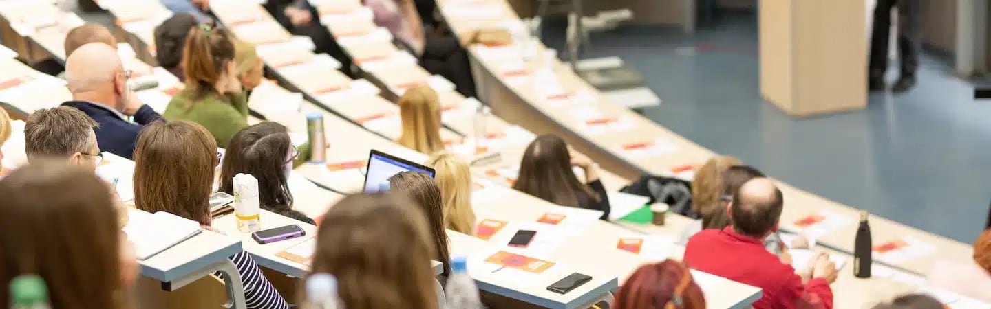 A diverse group of people attentively listening to a speaker in a lecture hall filled with long desks and notepads.