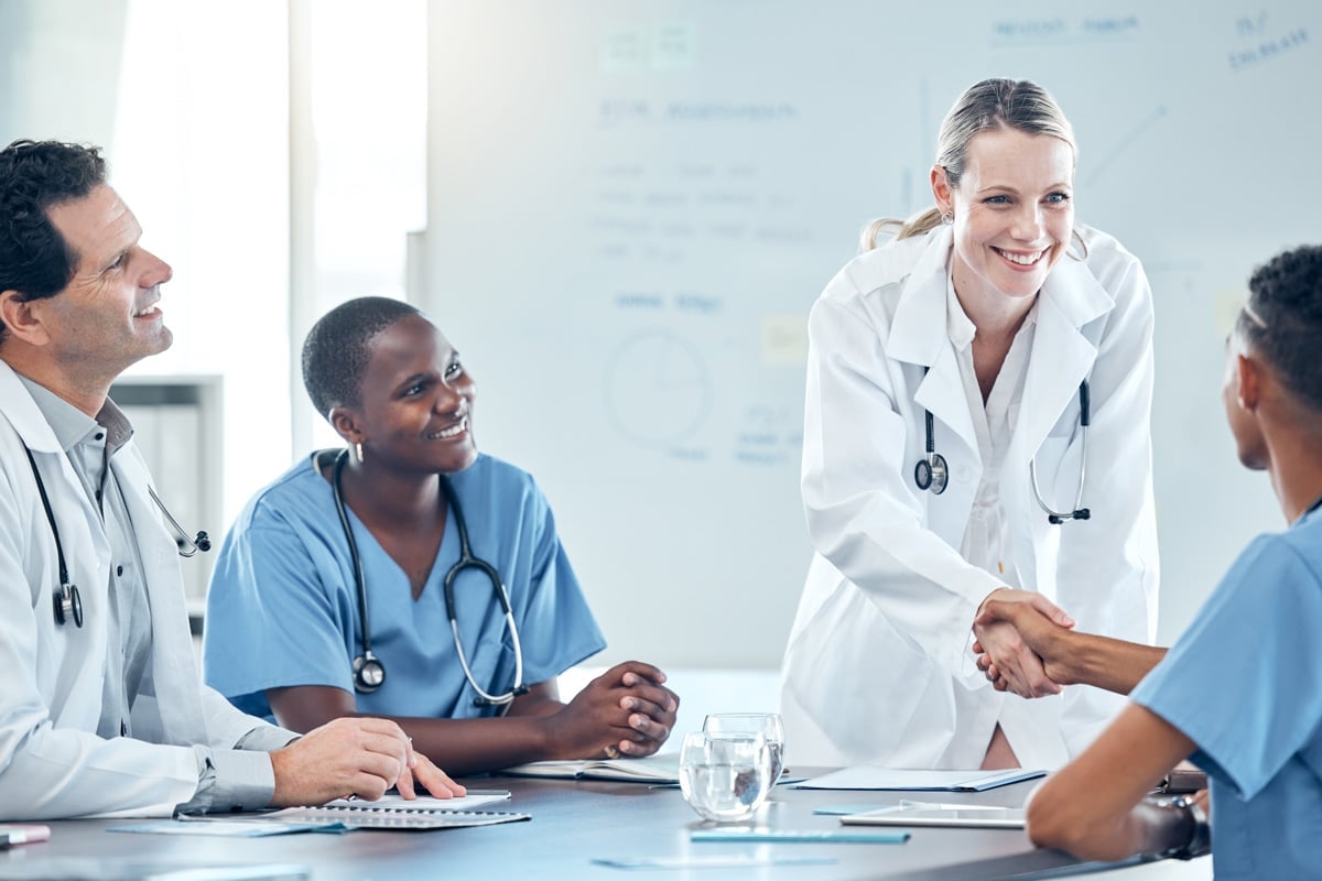 Healthcare professionals smiling and engaging in a meeting; two people in lab coats, two in scrubs, one shaking hands.