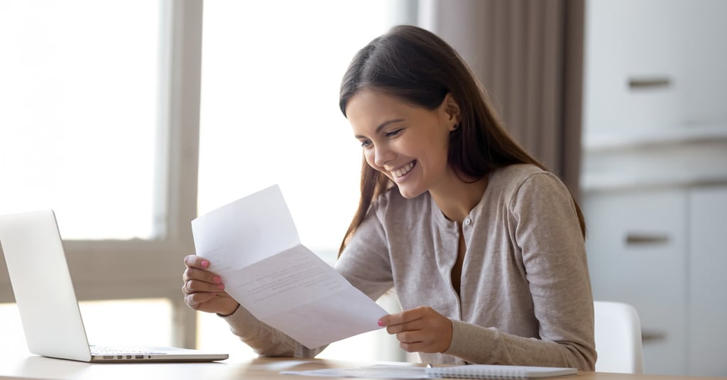 A smiling woman sits at a desk, looking at a letter in her hands about Financial Aid, with an open laptop and some papers in front of her, appearing happy and pleasantly surprised.