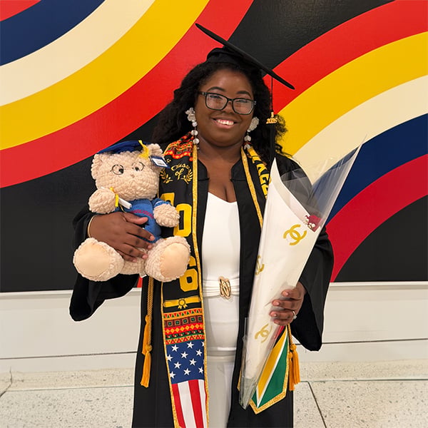 A smiling graduate, Keziah Williams-Daniels, in a cap and gown holds a bouquet and a teddy bear, standing in front of a colorful background with curved red, yellow, blue, and white stripes—proudly celebrating her achievement in AAS Paralegal Studies.