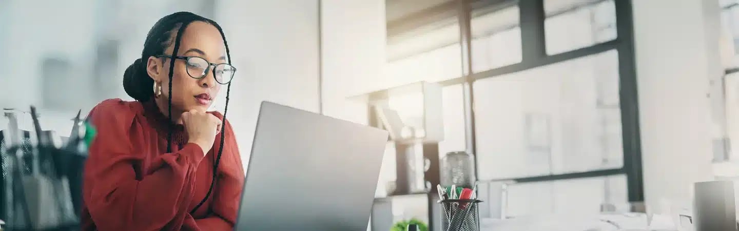 Woman with glasses working on a laptop at a desk in a bright office setting.