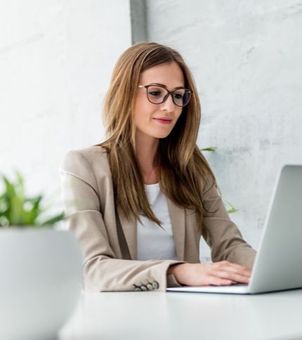 A Social Media Manager with long brown hair and glasses, wearing a beige blazer and white shirt, works on a laptop at a white desk. A potted plant is visible in the foreground.