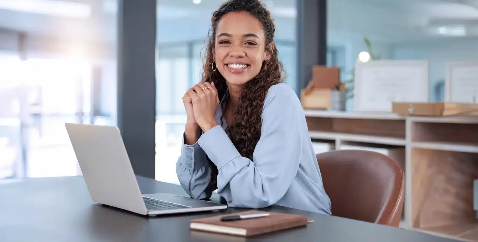 Smiling person sits at desk with open laptop in a modern office setting with shelves and large windows in the background.