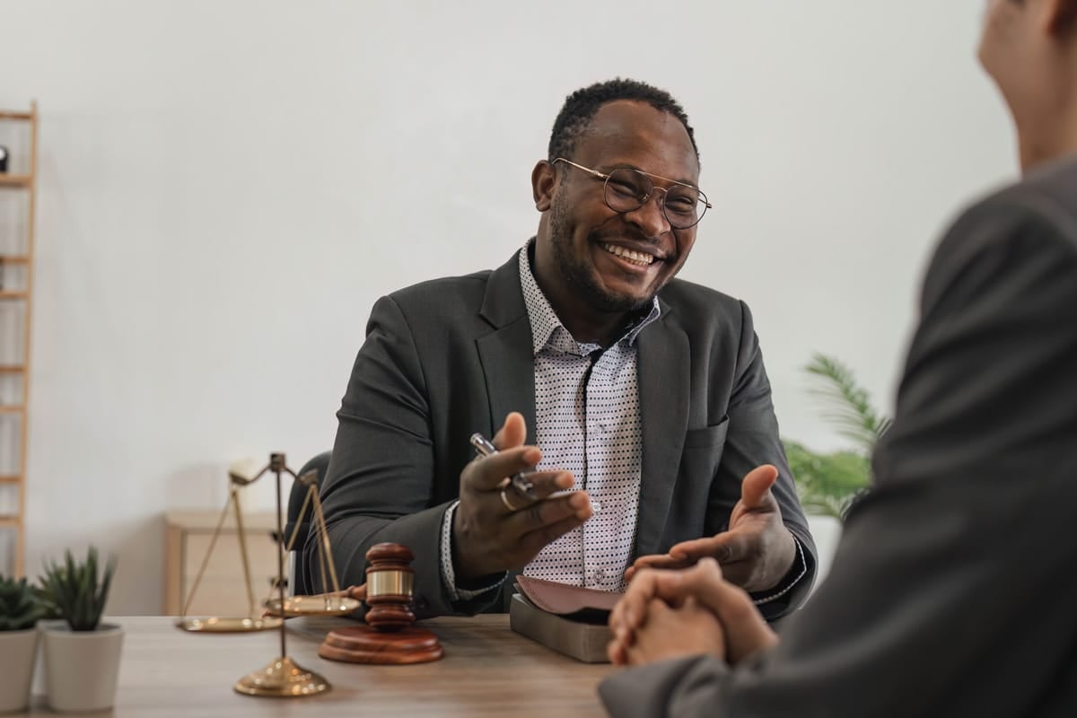 Smiling man in a suit sits at a desk with legal items, talking to another person with hands clasped.