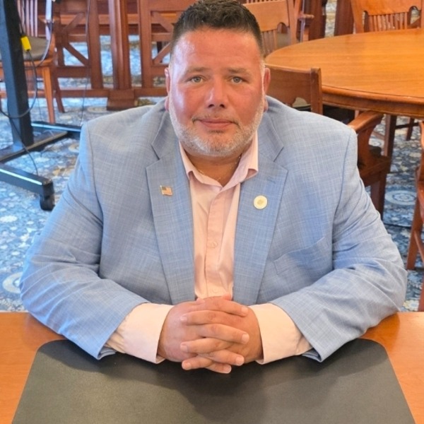 A man, identified as Kevin Meaker, in a light blue suit and pink shirt sits at a desk with his hands clasped, in a room with wooden tables and chairs.