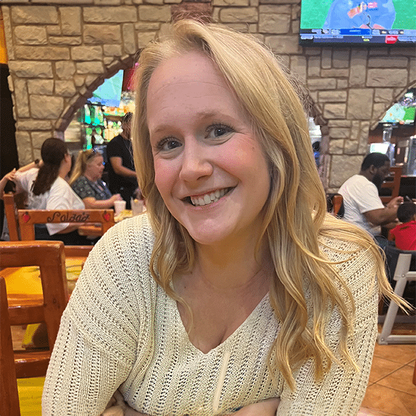 Smiling woman with long blonde hair and wearing a white sweater, seated in a restaurant with stone walls in the background.
