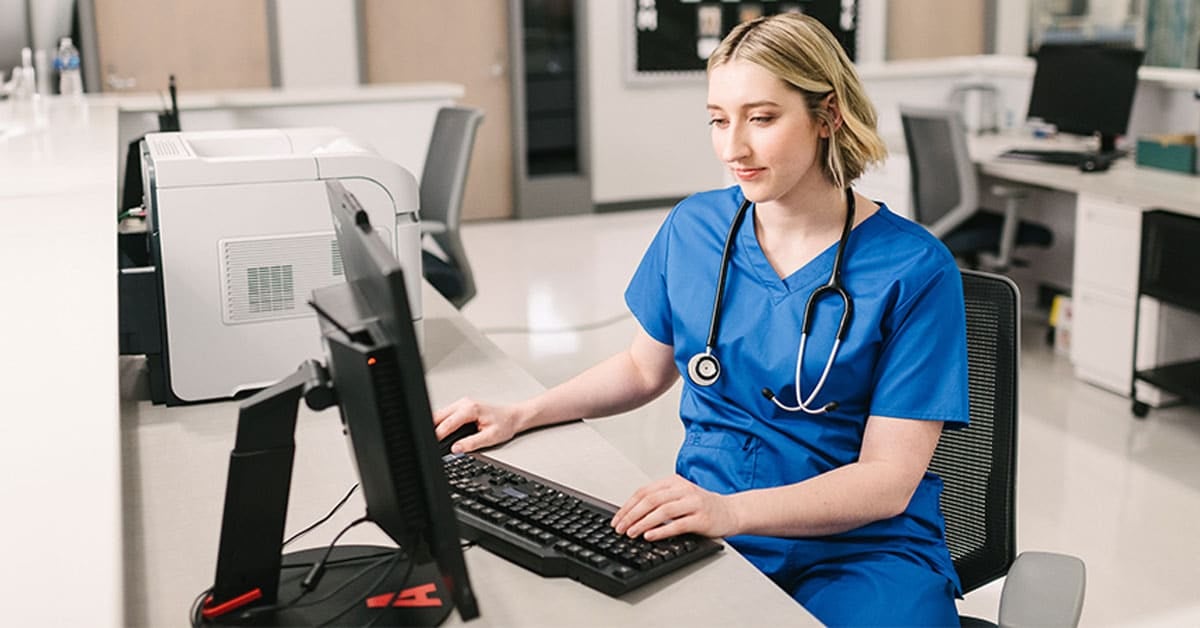 A healthcare professional in blue scrubs working on a computer in a medical office, wearing a stethoscope.