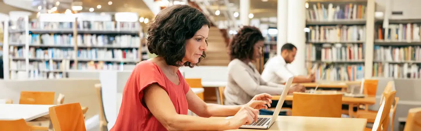 People working on laptops at a library table, with shelves of books and a staircase in the background.