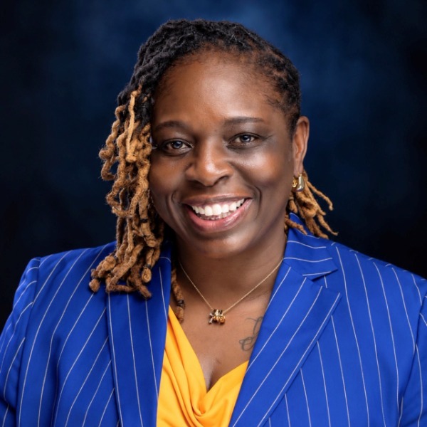 A smiling woman with locs wearing a blue pinstripe blazer over a yellow top.