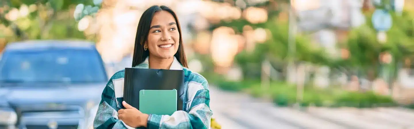 Smiling person holding notebooks and wearing a green plaid shirt, standing outside with a car and greenery in the background.