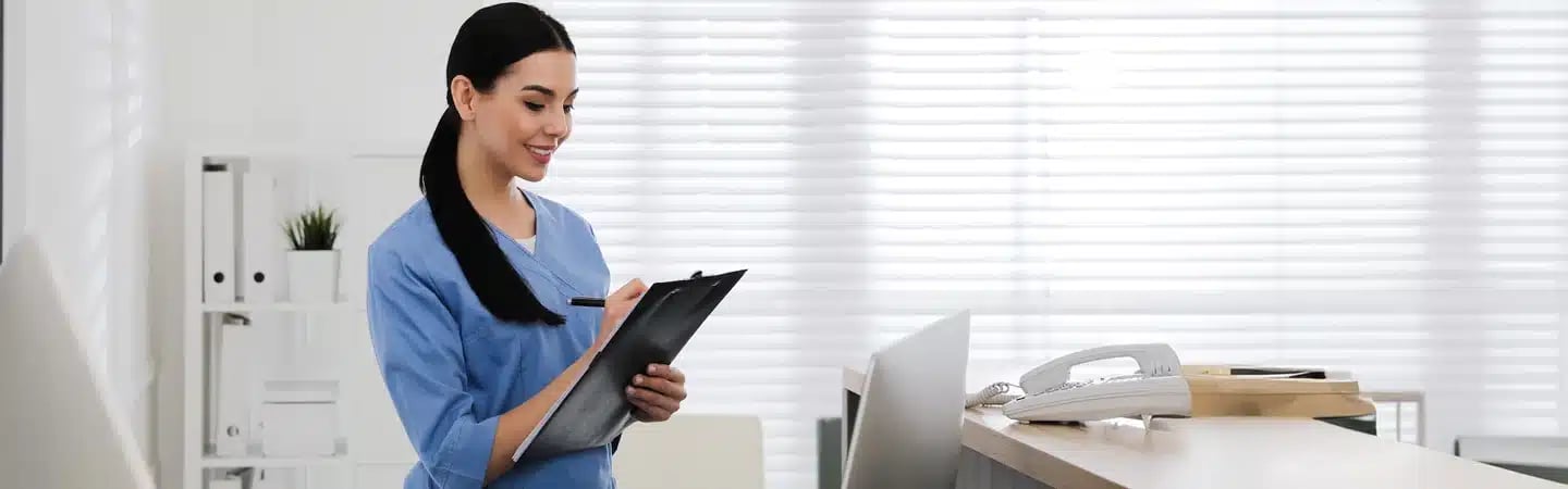 A nurse in blue scrubs smiles while writing on a clipboard at a reception desk. A phone and laptop are on the desk.