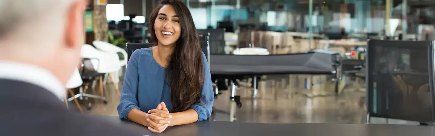 A woman smiles during a meeting at a sleek, modern office with glass walls and open workspace in the background.