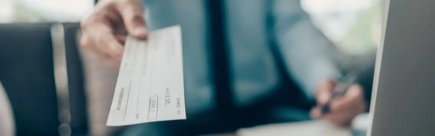 A hand holds out a check while a person in business attire writes at a desk in the background.