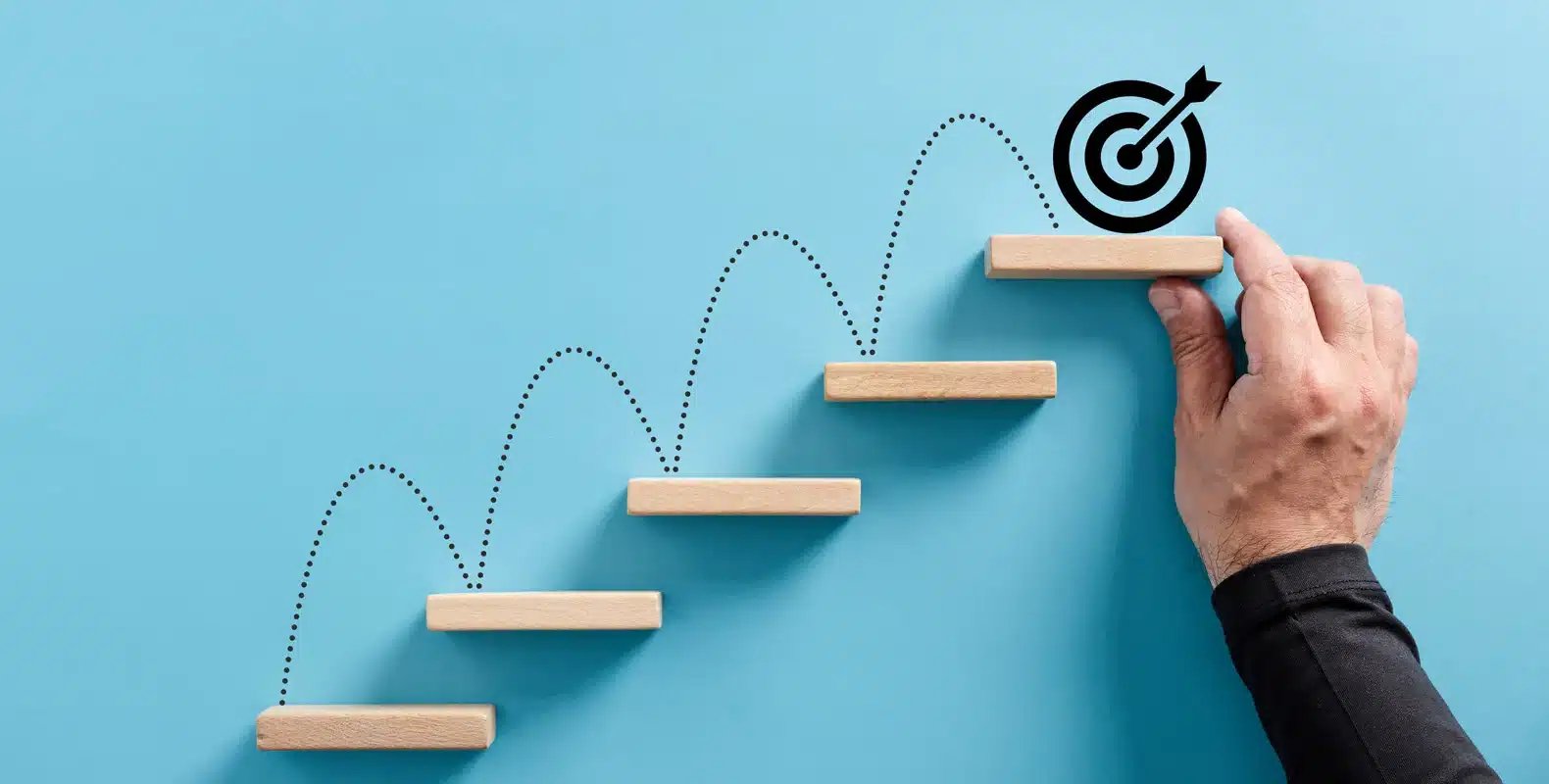 A hand arranges wooden steps leading to a target symbol, representing progression and reaching goals against a blue background.