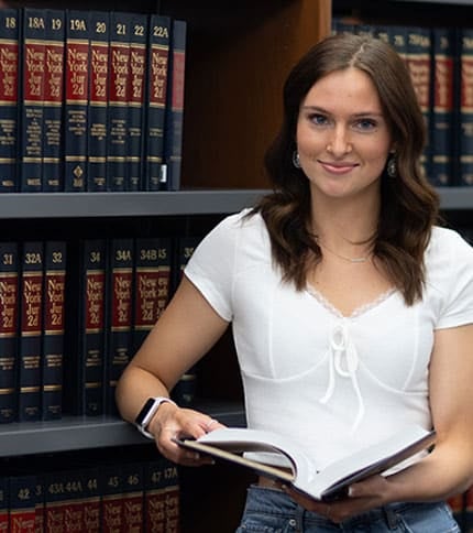 A person with long brown hair stands in front of shelves filled with New York legal books, holding an open book. They're wearing a white top and jeans, and smiling at the camera.