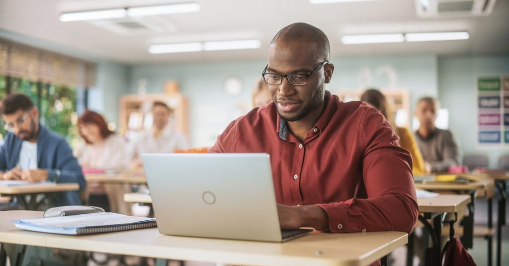 A man wearing glasses and a red shirt works on a laptop at a desk in a classroom, possibly researching Financial Aid options, with other adults seated and working in the background.