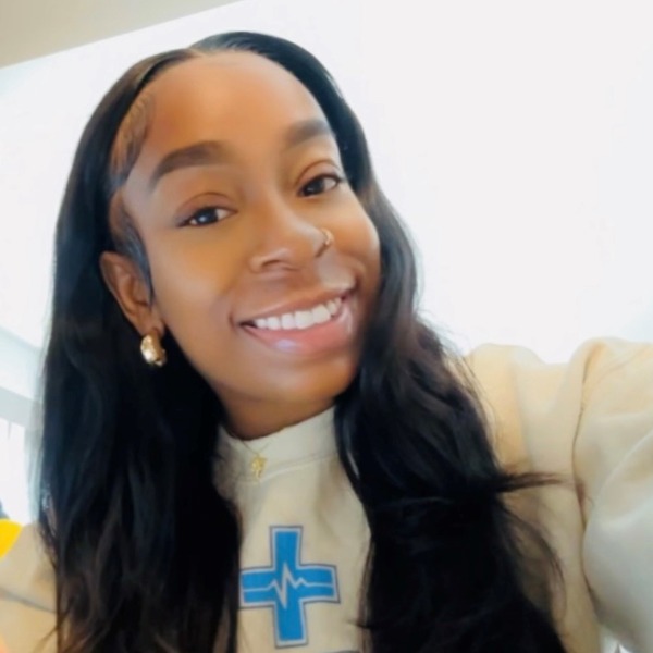 Woman with long black hair smiling, wearing a light sweatshirt with a blue medical symbol, gold earrings, and a nose ring, in a brightly lit room—Henrietta is an AAS Nursing student.
