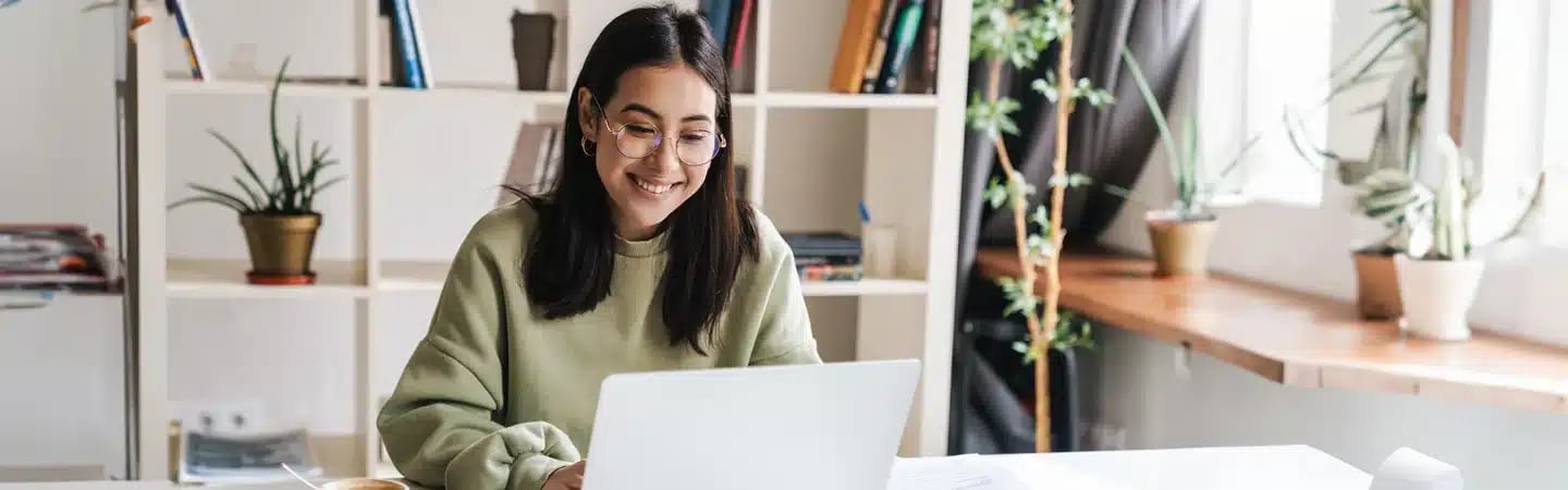 Woman with glasses in a green sweater smiling while working on a laptop at a bright, plant-filled workspace.