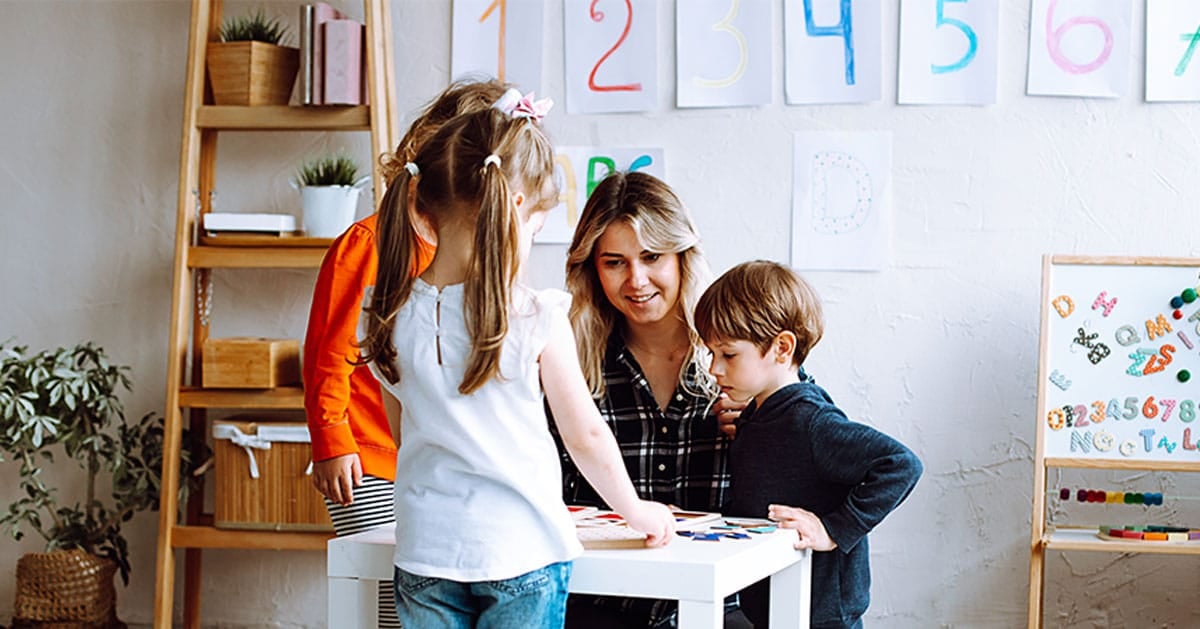 Woman with three young children engaged in an activity around a white table in a classroom with colorful numbers on the wall.
