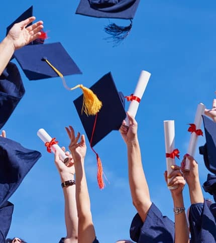 Graduates in blue gowns throw their caps into the air while holding diplomas with red ribbons, celebrating against a clear blue sky.