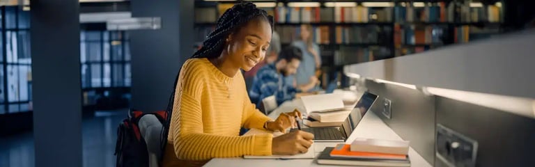 A person in a yellow sweater studies in a library, writing in a notebook and using a laptop.