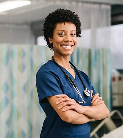 A healthcare professional in blue scrubs is smiling with arms crossed in a medical facility. A stethoscope hangs around their neck. The background features patterned privacy curtains and medical equipment.