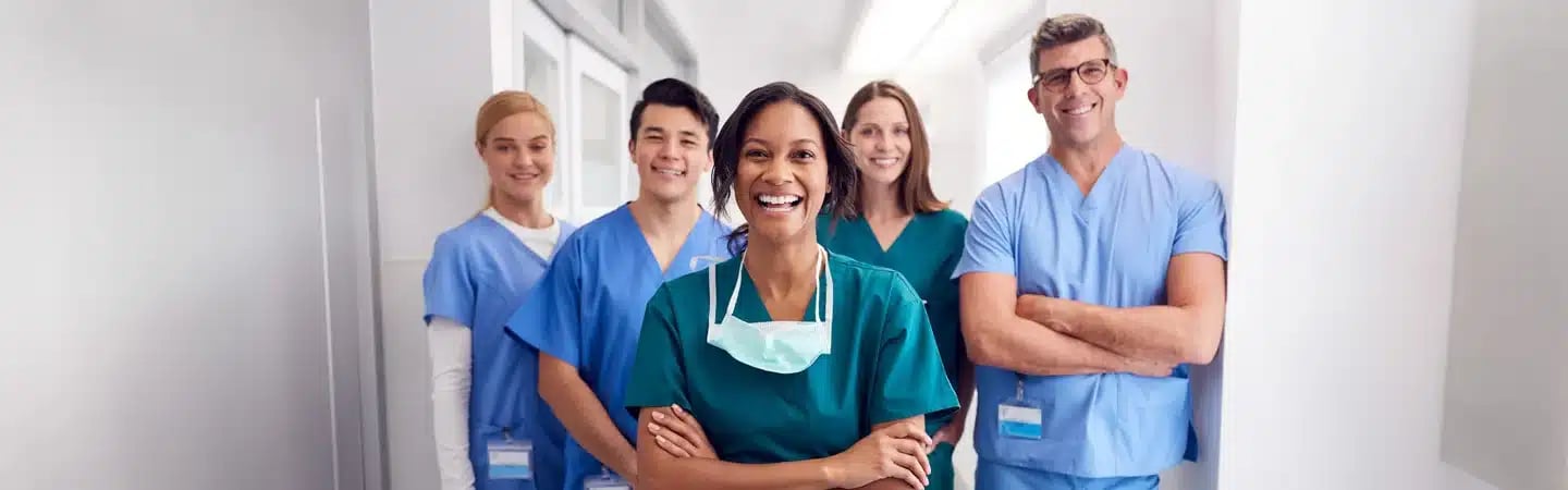 A diverse group of healthcare professionals in scrubs smiling and standing in a bright hallway.