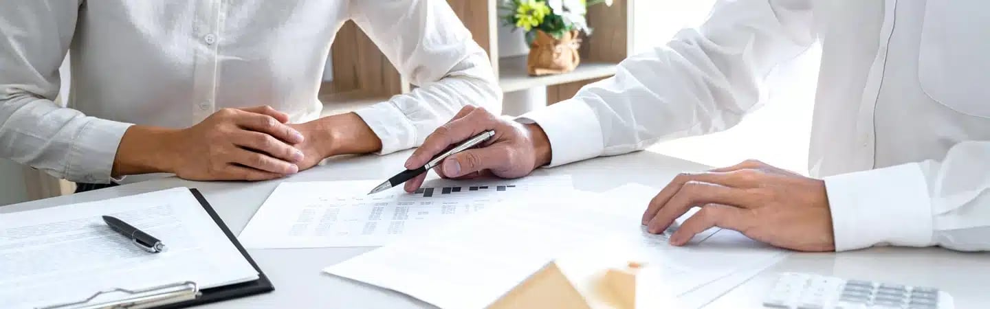 Two people in white shirts discussing documents at a table, one holding a pen pointing to a paper.