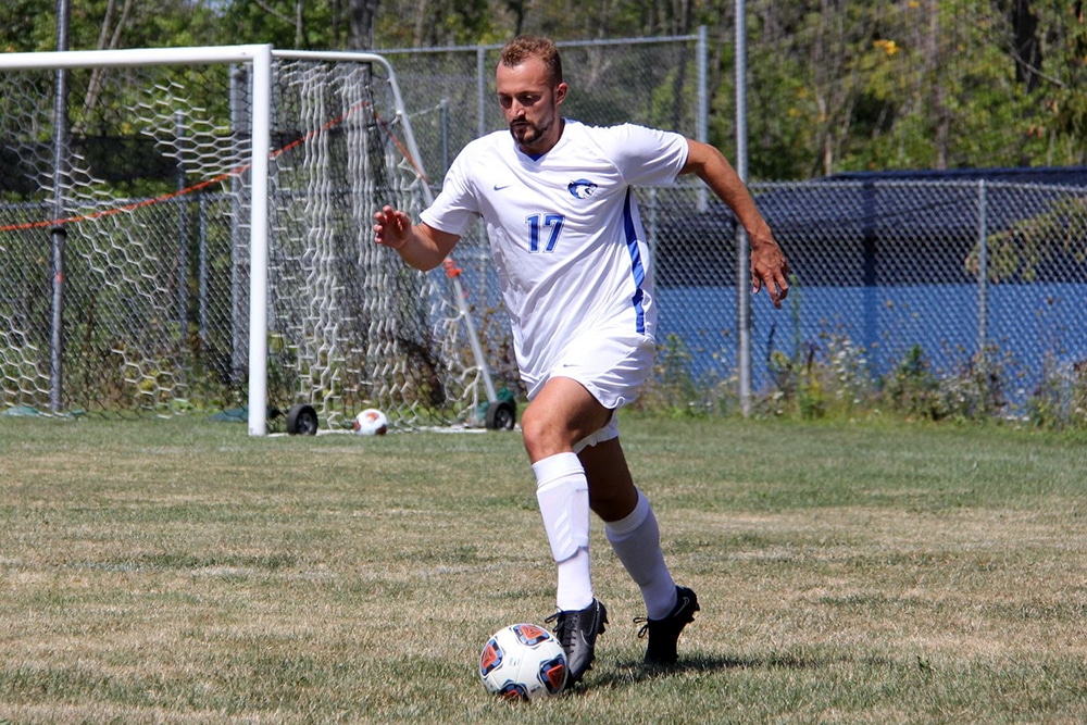 A soccer player in a white uniform and the number 17 dribbles the ball on a grassy field in daylight.