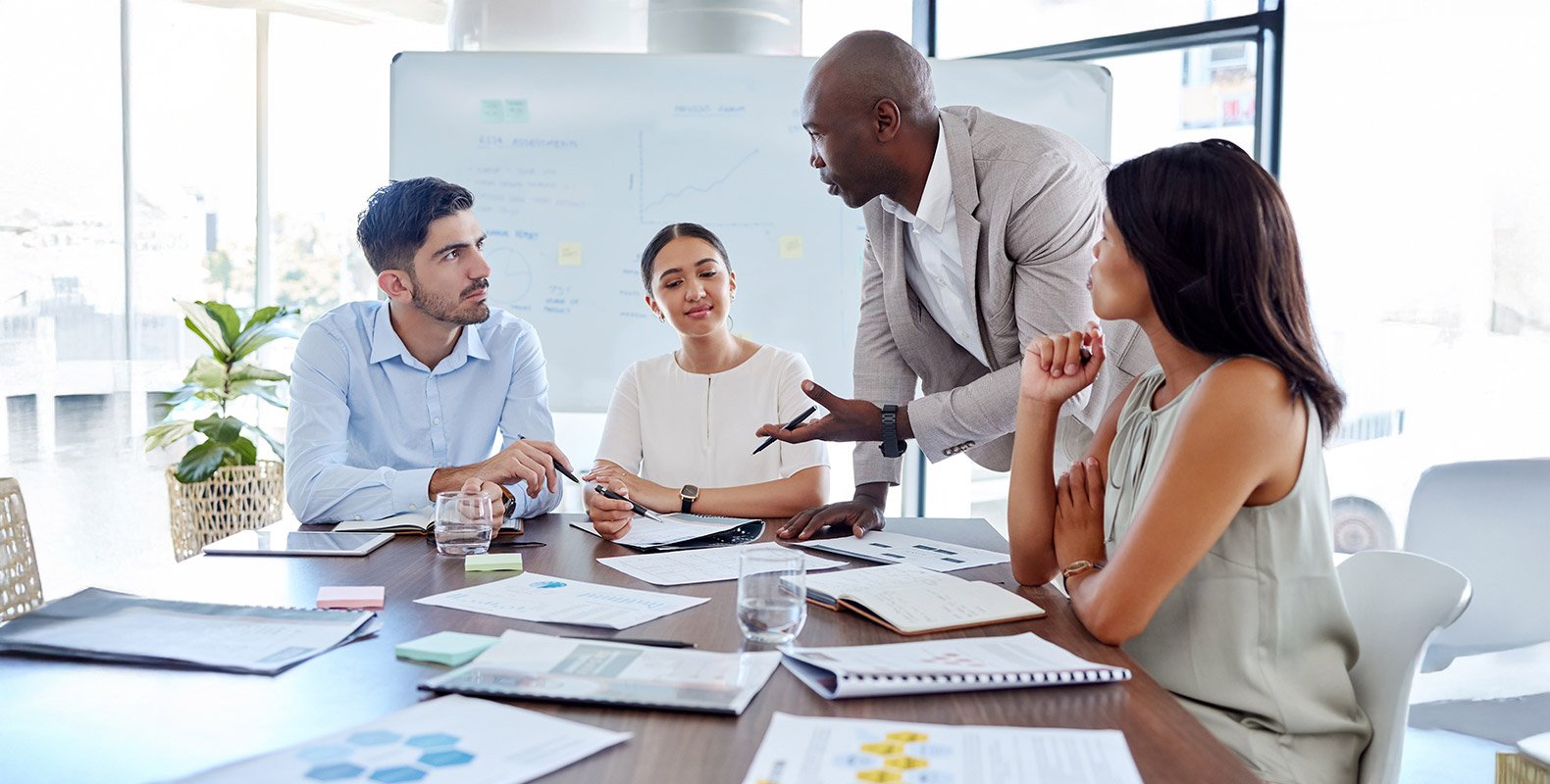 Four people in business attire sit around a table with papers and laptops, engaged in discussion. One person stands, gesturing while speaking, as the others listen attentively in a modern office setting.