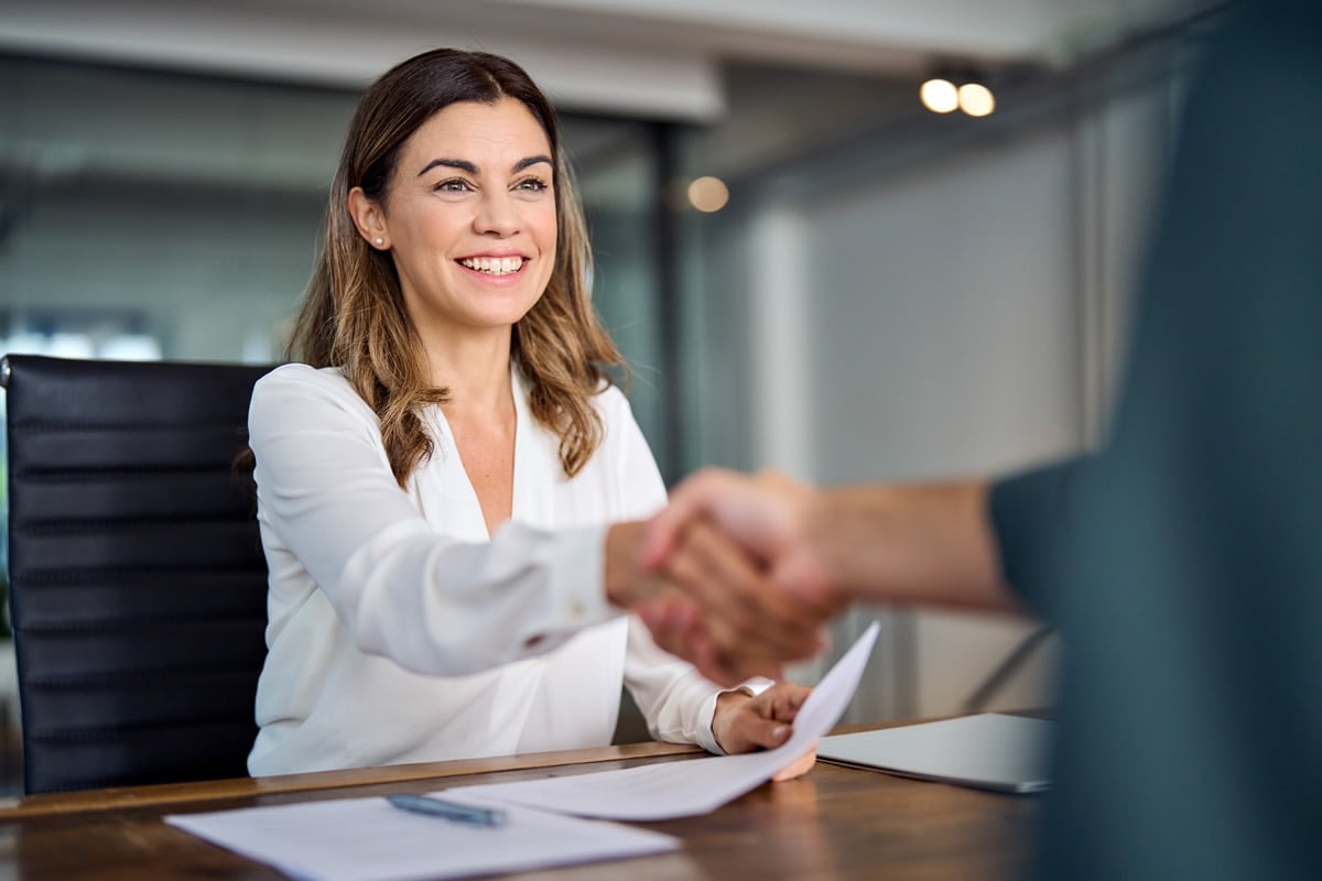 Woman in white blouse smiling and shaking hands with someone across a desk, holding a document.