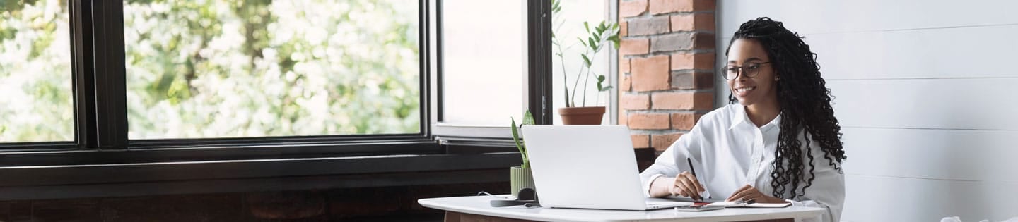 A person with glasses working on a laptop at a desk, with large windows and plants in the background.