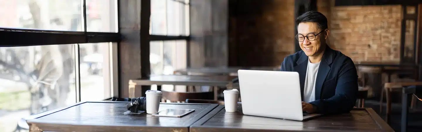 A person in glasses using a laptop at a café table with a coffee cup and tablet, looking at the screen and smiling.