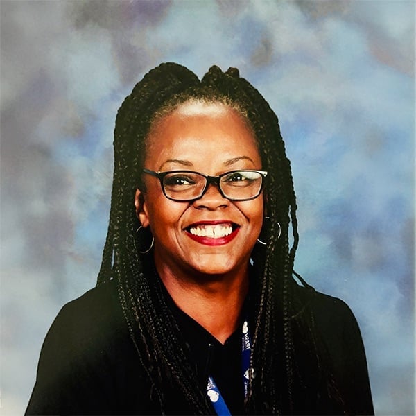 A woman with long braided hair, glasses, and hoop earrings is smiling widely. She is wearing a black top and a lanyard, with a soft blue and gray background behind her.