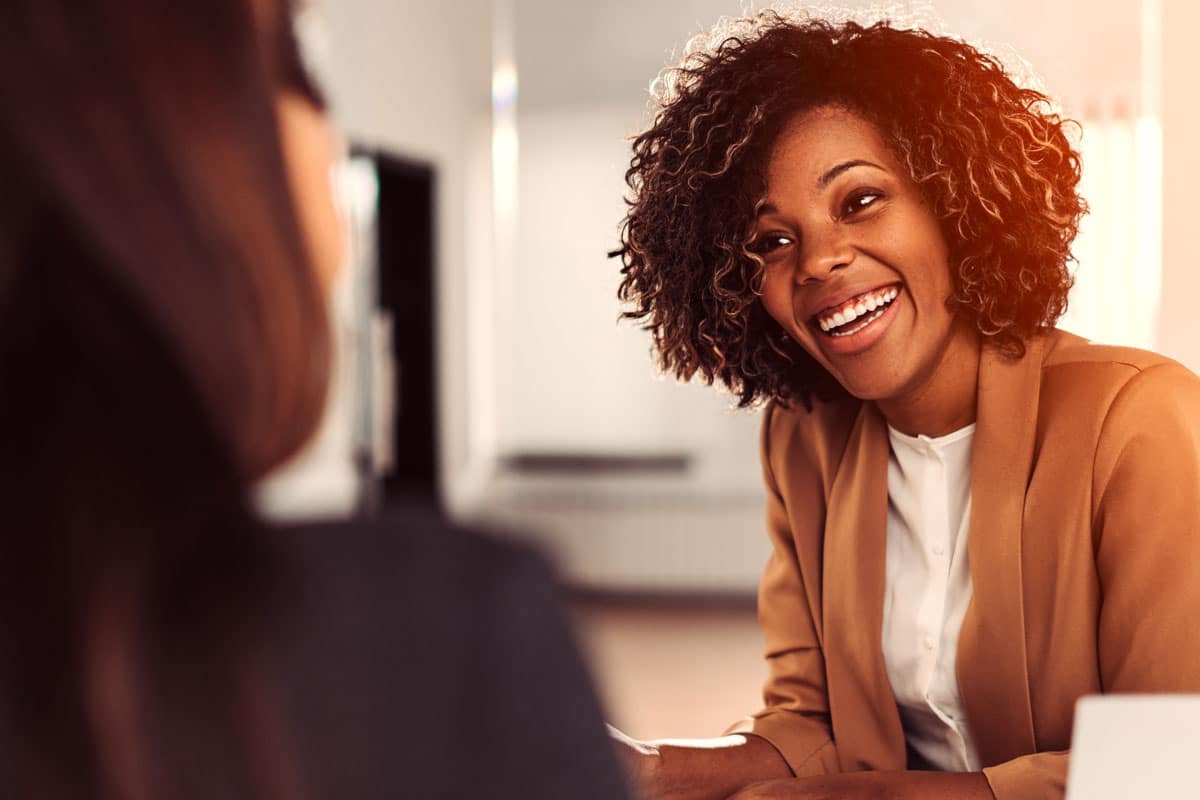 A woman with curly hair, wearing a tan blazer, smiles warmly during a conversation with another person who is out of focus in the foreground. They appear to be in a professional or office setting.
