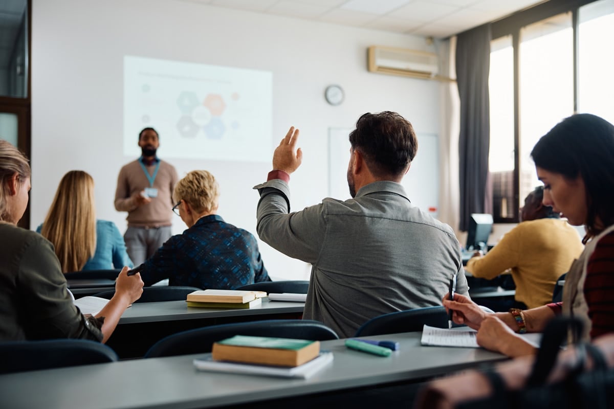A person raises their hand in a classroom setting during a lecture. Other attendees are seated and facing the presenter.