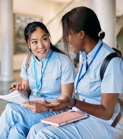 Two women in blue uniforms sit and converse while holding notebooks, their ID lanyards reflecting a commitment to excellence. They appear engaged in discussing online healthcare degrees, set against a bright indoor background.