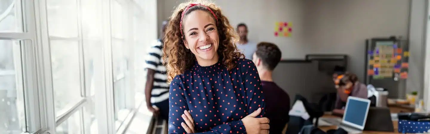 Smiling woman in a polka dot shirt stands with arms crossed in a bright office with people working in the background.