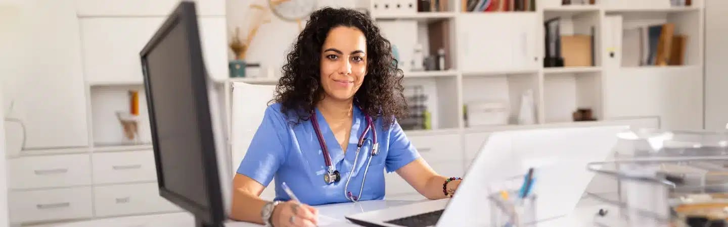 A healthcare professional in scrubs, with a stethoscope around her neck, sits at a desk using a computer.