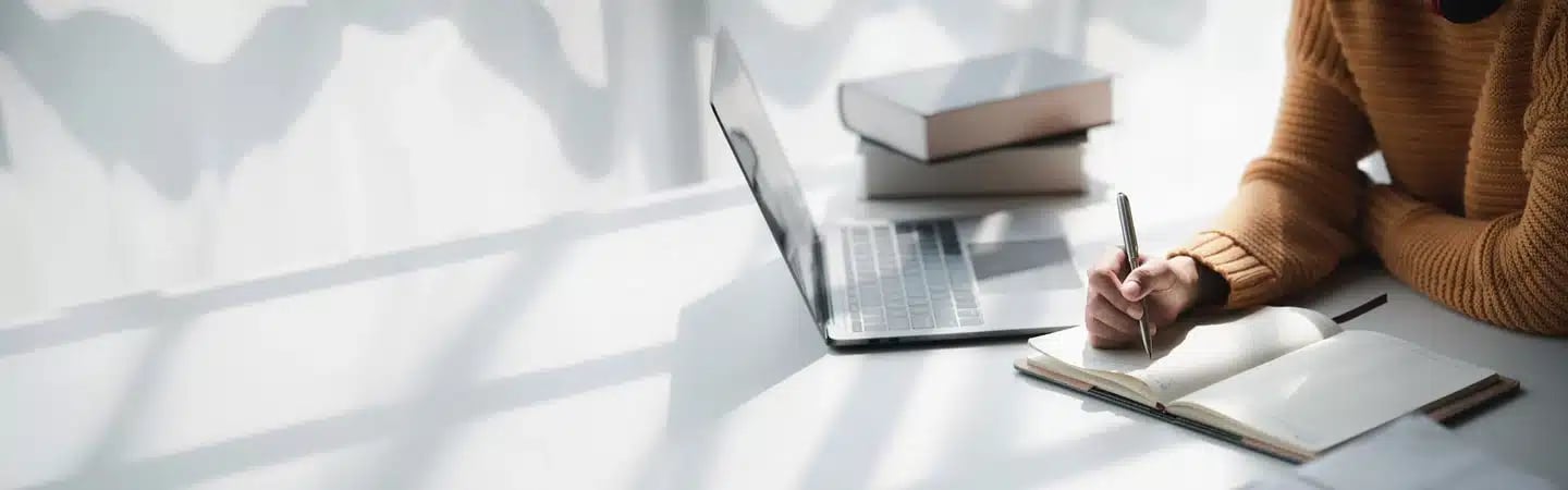 Person in a yellow sweater writing in a notebook beside a laptop and a stack of books on a sunlit desk.