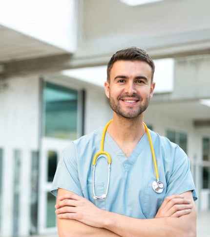 A young male healthcare professional in light blue scrubs with a yellow stethoscope around his neck stands outdoors, smiling with his arms crossed in front of a modern medical building.