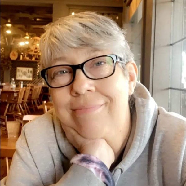 A person with gray hair and glasses smiles, resting their chin on their hand, seated in a cozy, warmly-lit room.