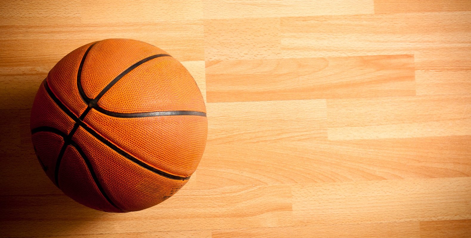 A basketball resting on a hardwood court, illuminated from the side.