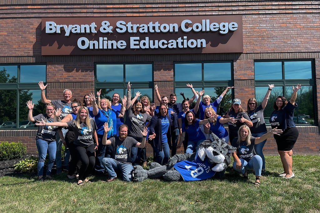 A group of people pose with raised arms in front of the Bryant & Stratton College Online Education building.