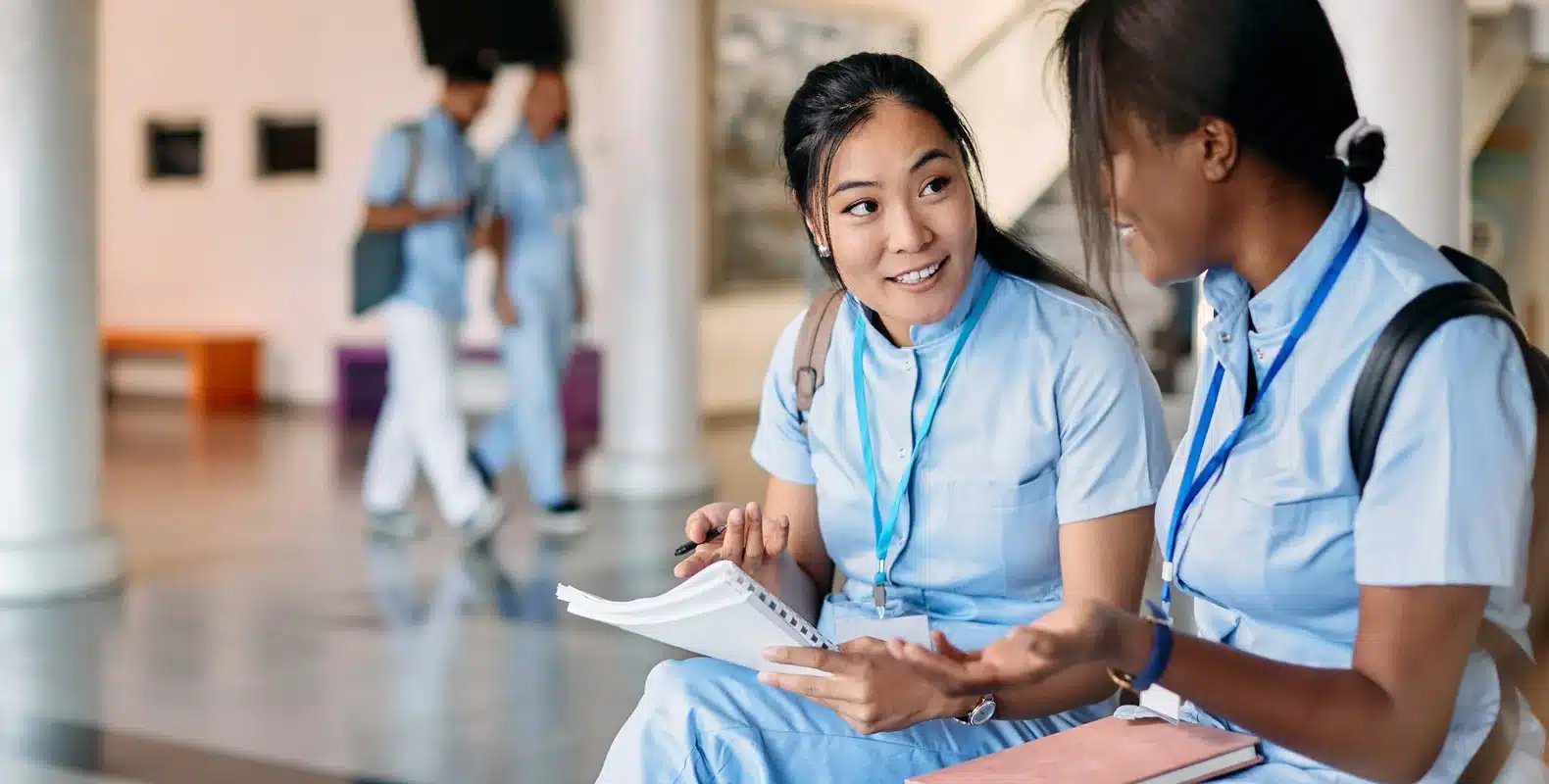 Two students in uniforms sit and talk, holding notebooks. Two other students are in the background, also in conversation.